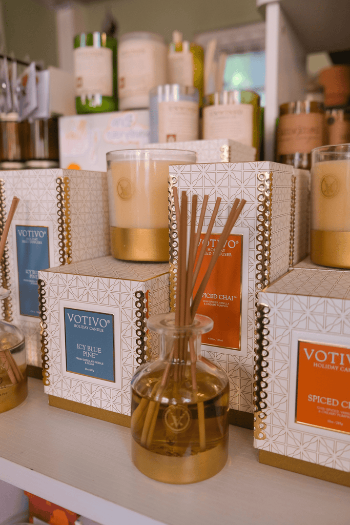 Shelves displaying Votivo candles and reed diffusers in elegant packaging. The products are primarily in blue and gold colors, including scents like Icy Blue Pine and Spiced Chai, with a warm, inviting atmosphere.