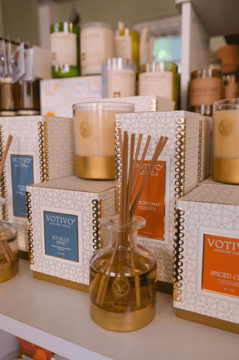 Shelves displaying Votivo candles and reed diffusers in elegant packaging. The products are primarily in blue and gold colors, including scents like Icy Blue Pine and Spiced Chai, with a warm, inviting atmosphere.