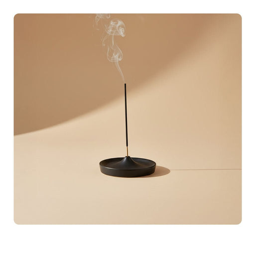 A thin incense stick burns in a minimalist black holder, releasing a gentle trail of smoke against a soft beige background.