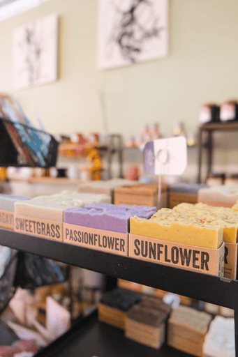 A shelf displays various handmade soaps labeled with names like Sweetgrass, Passionflower, and Sunflower. The background shows wooden shelves with more soaps and items in a cozy store setting.