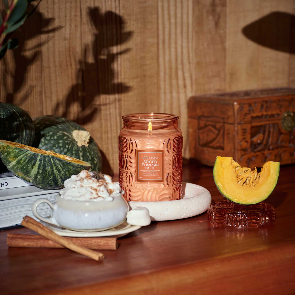 A lit Voluspa Spiced Pumpkin Latte Large Jar 18oz Candle made from a coconut wax blend sits on a table with a frothy drink, whipped cream, cinnamon, books, green and orange pumpkins, and a carved wooden box.
