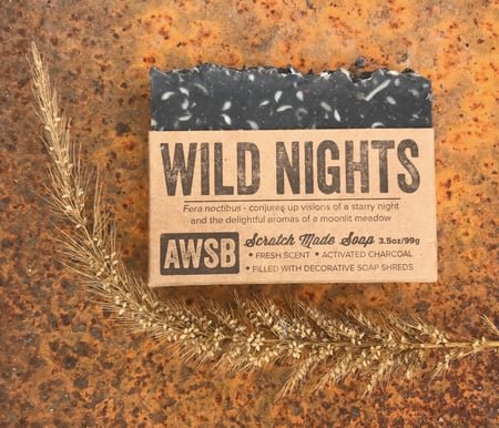 A Wild Soap Bar - Wild Nights by A Wild Soap Bar rests on rustic terrain with dried grass. Featuring essential oils, a fresh scent, activated charcoal, and decorative soap shreds, this handcrafted bar is ideal for zero-waste enthusiasts.