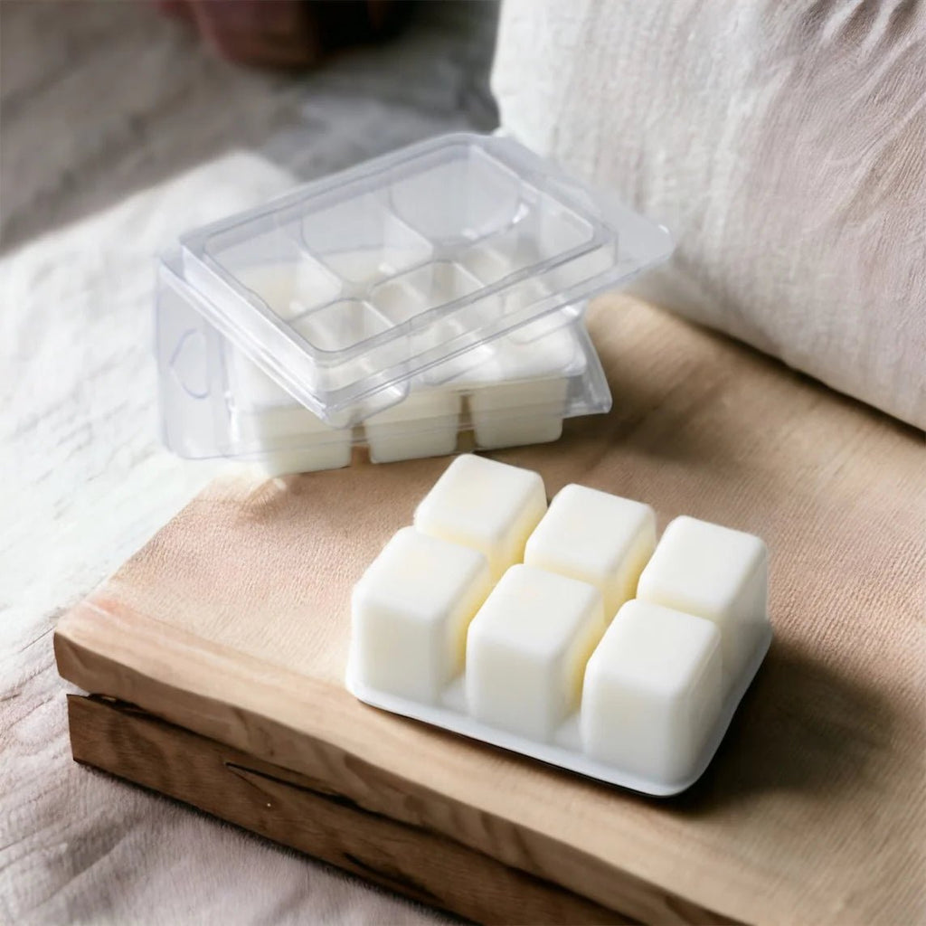 A plastic container with a hinged lid holds six Allusions - Morning Buzz Soy Beeswax Melts on a wooden surface. Another similar container sits nearby with its lid open, revealing more melts. A plush, light-toned fabric is visible in the background.