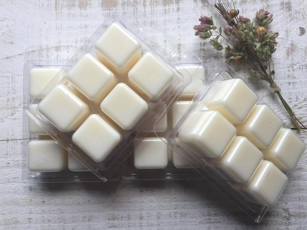 Three packs of Allusions - Morning Buzz Soy Beeswax Melts 3oz are artfully arranged on a textured wooden surface with a small bouquet of dried flowers. The melts, displayed in rectangular plastic molds with six cubes each, are hand-poured in the USA.