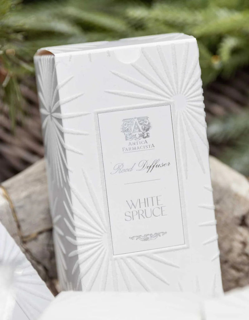 The Antica Farmacista 320ml White Spruce Reed Diffuser offers an Evergreen Fragrance, packaged in a white box with embossed starburst patterns and text, set against blurred greenery.