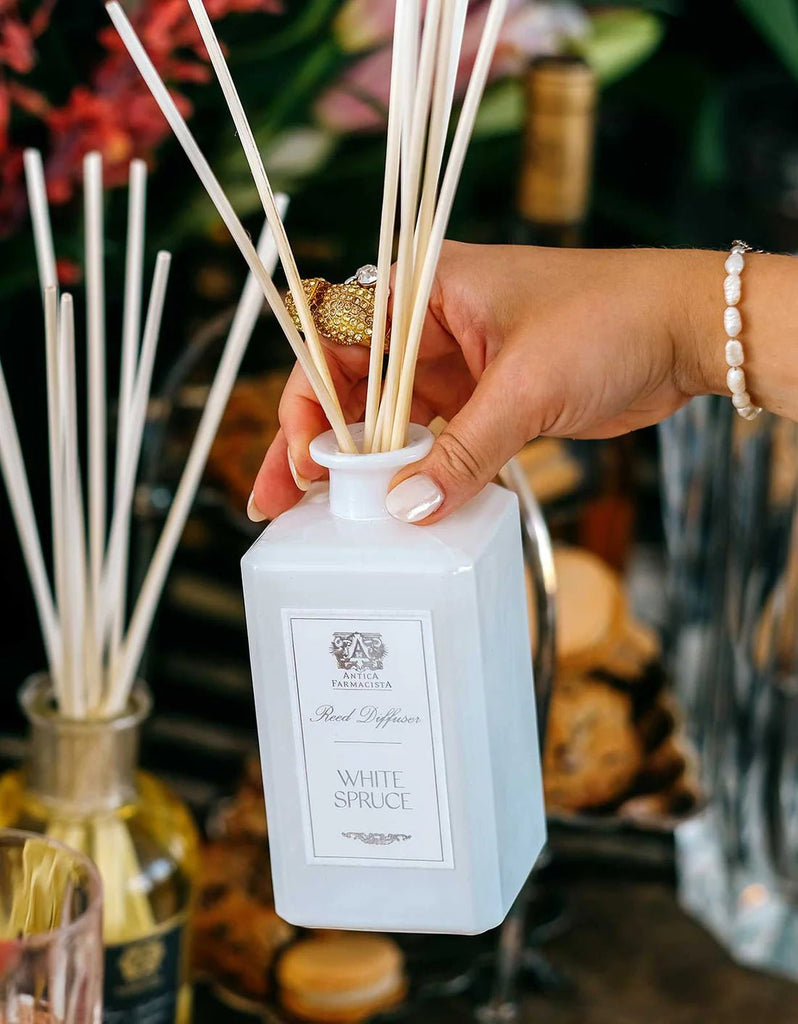 A bracelet-clad hand elegantly inserts wooden reeds into an Antica Farmacista White Spruce Reed Diffuser, set amidst apothecary-inspired bottles, filling the room with an enchanting evergreen fragrance.