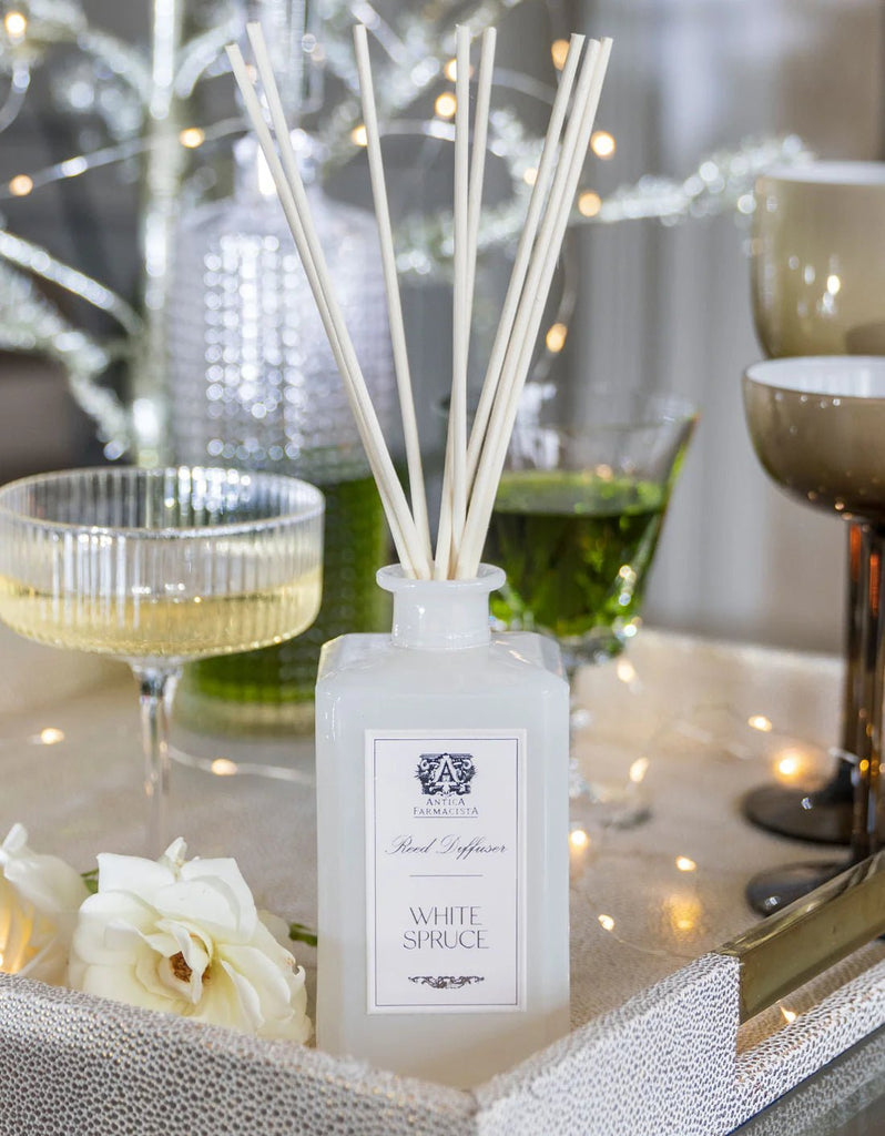 An Antica Farmacista - 320ml White Spruce Reed Diffuser sits on a tray with candles and sparkling drinks, accompanied by a white rose. A lit decorative tree enhances the elegant scene with evergreen fragrance.