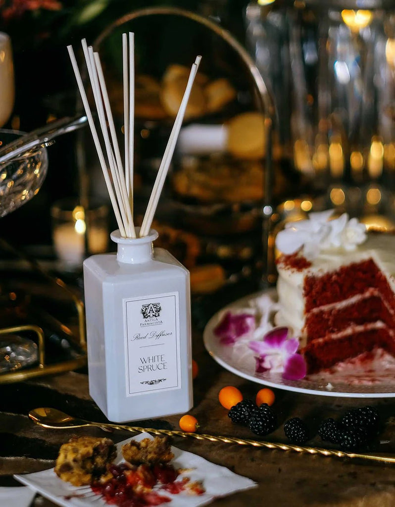 Antica Farmacistas 320ml White Spruce Reed Diffuser, in an apothecary-style bottle, enriches a dessert table with its evergreen scent. Nearby, a red velvet cake with orchids joins berries and a pie under warm lighting.