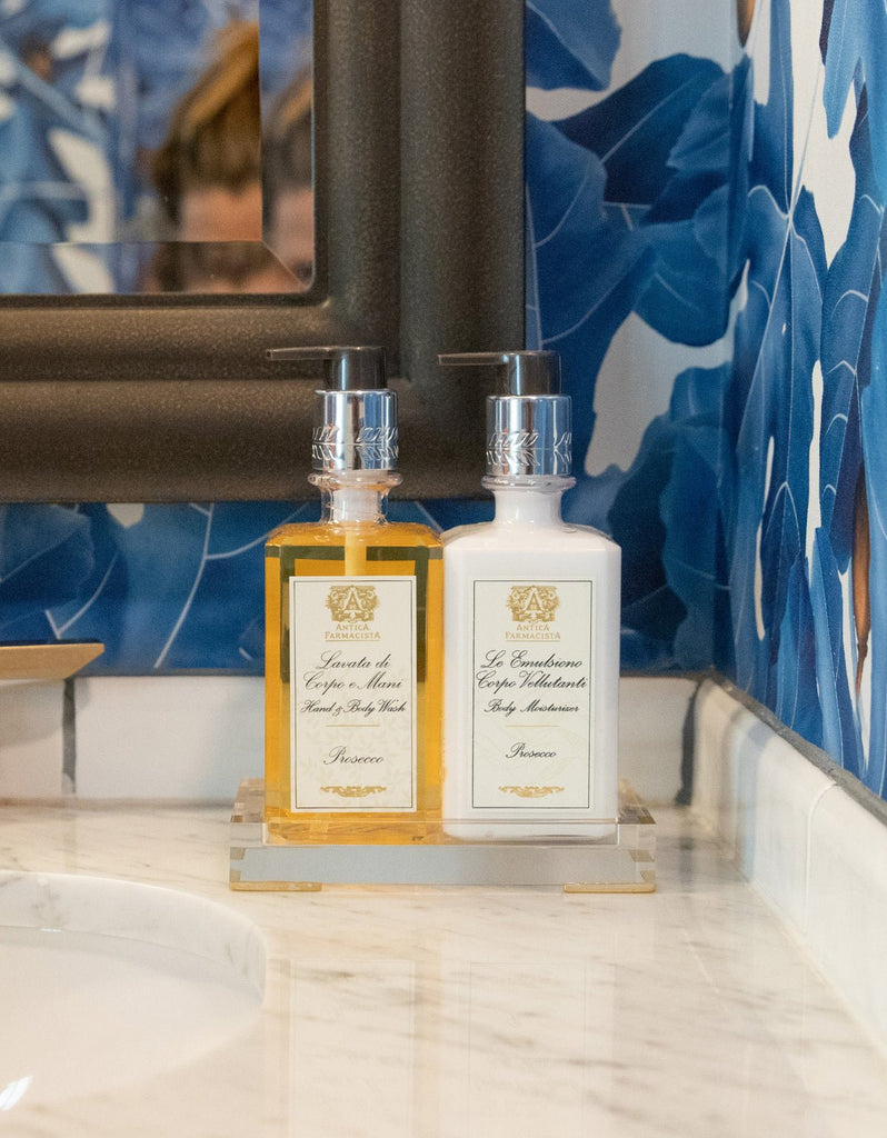 An Antica Farmacista Acrylic Tray holds 10oz amber and white bottles of Hand Wash and Body Moisturizer on a marble countertop. The wall showcases a bold blue and white floral pattern, with a rectangular mirror partly visible above the tray.