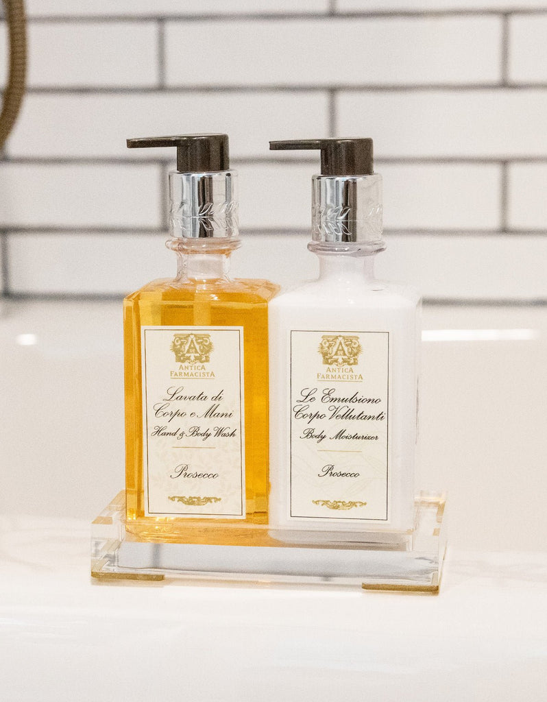 Two elegant Antica Farmacista bottles rest on an acrylic tray against white tiles. The left holds orange Hand Wash, the right contains white Body Moisturizer. Both feature silver pumps, adding sophistication to any bathroom décor.