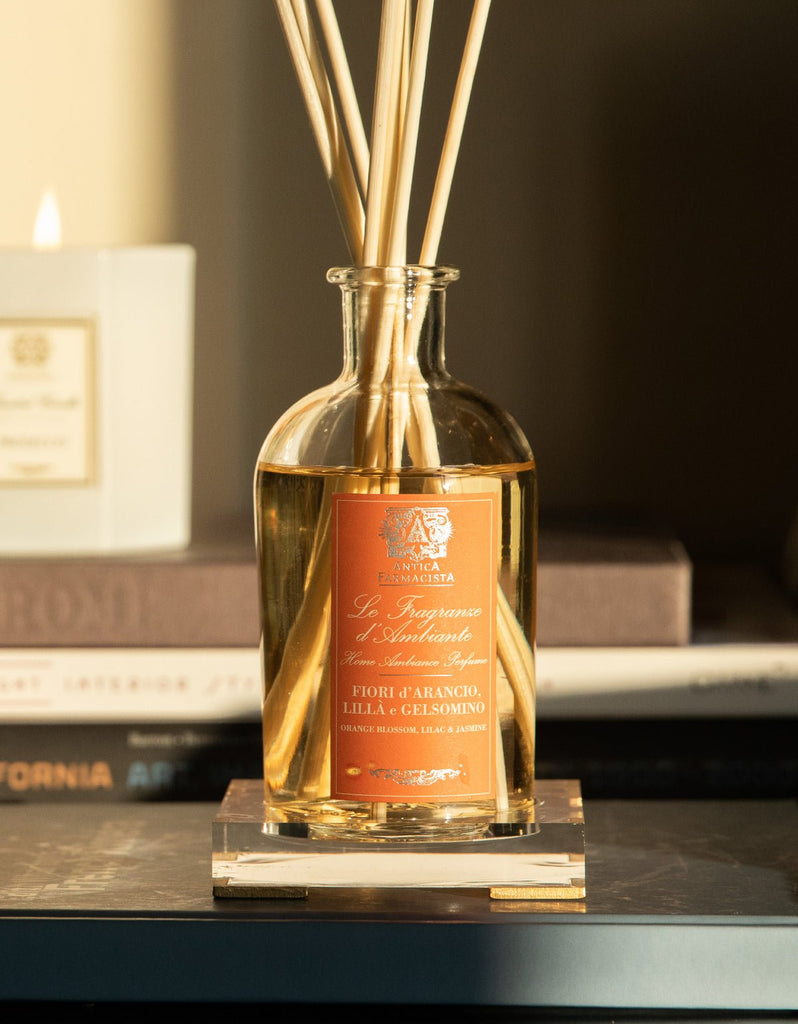 A 250ml Antica Farmacista reed diffuser with wooden reeds is elegantly displayed on a small stand. The label shows Fior dArancio, Lilla e Gelsomino. Stacked books and a lit candle accompany it on an acrylic tray, creating a cozy atmosphere under warm lighting.