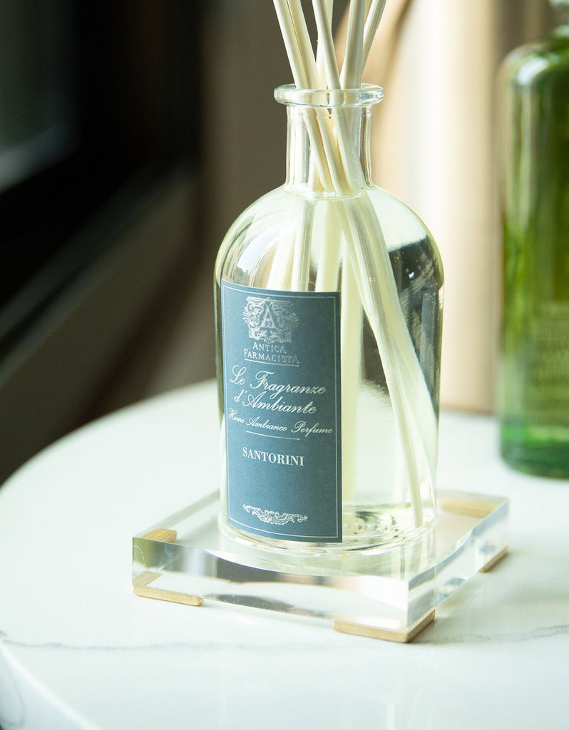 A clear glass diffuser bottle labeled Le Fragranze dAmbiente Santorini stands elegantly on a marble surface, part of the Antica Farmacista 250ml Diffuser collection. White reed sticks fan out from the top, with a green bottle blurred behind.