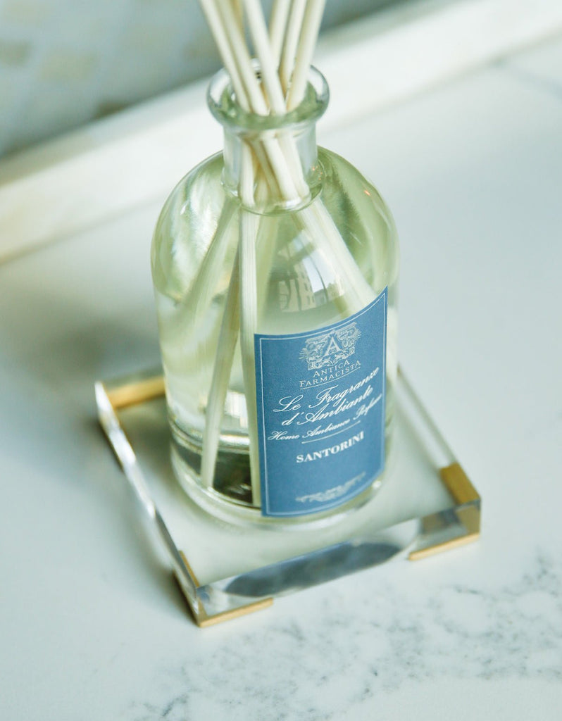 A sleek Antica Farmacista glass bottle with a blue Santorini label rests elegantly on an acrylic tray designed for a 250ml diffuser. The transparent scented oil stands out against the light marbled background.