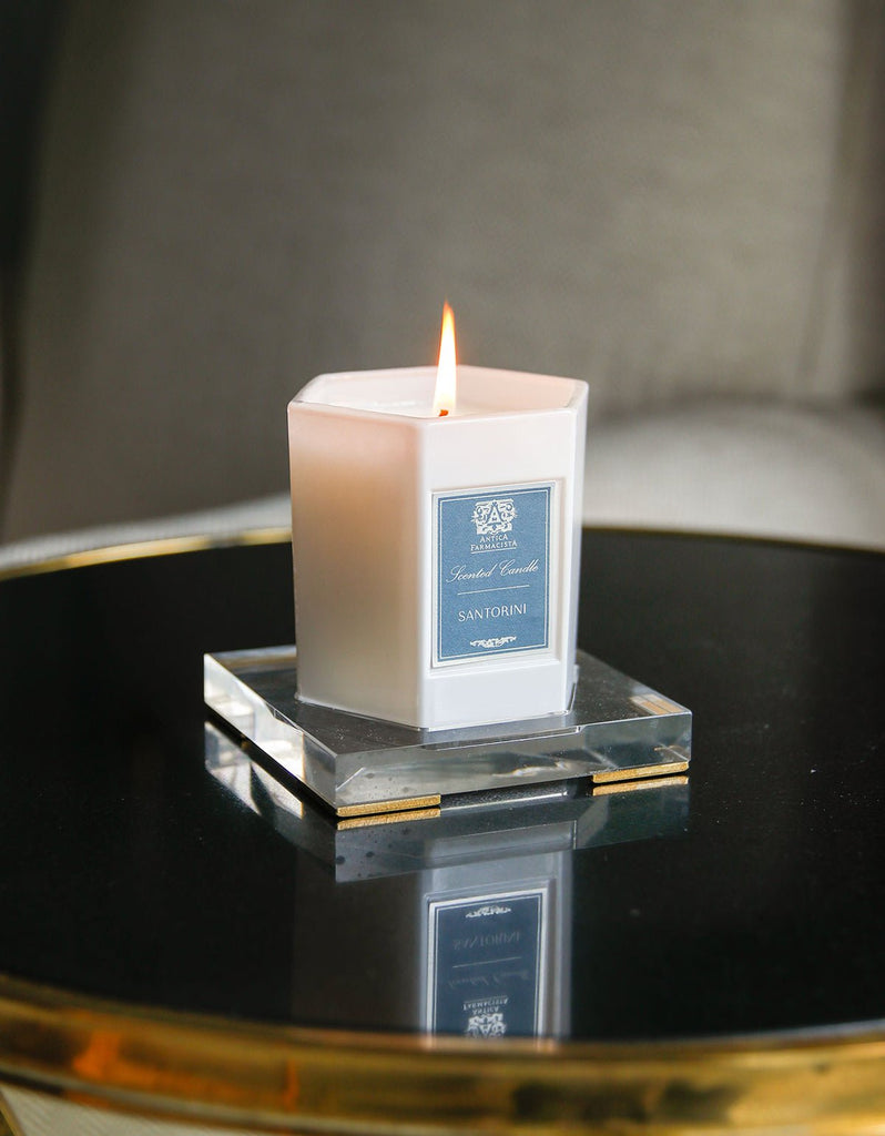 A lit 9oz candle by Antica Farmacista, labeled Santorini, sits in a hexagonal glass holder on an acrylic tray from Antica Farmacista. It rests elegantly on a reflective black surface against a mysteriously blurred gray background.