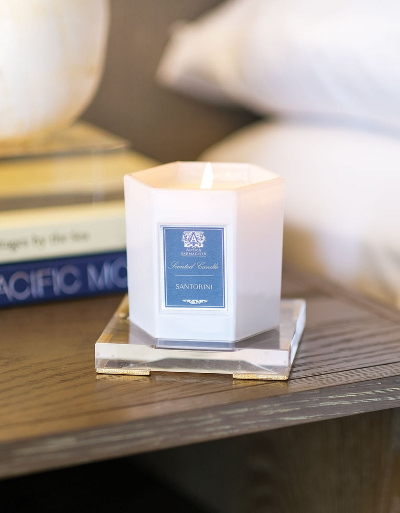 A lit 9oz candle labeled Santorini glows warmly on a hexagonal coaster. Nestled beside stacked books, it rests on the Antica Farmacista - Acrylic Tray for 9oz Candle, adding elegance to the wooden surface.