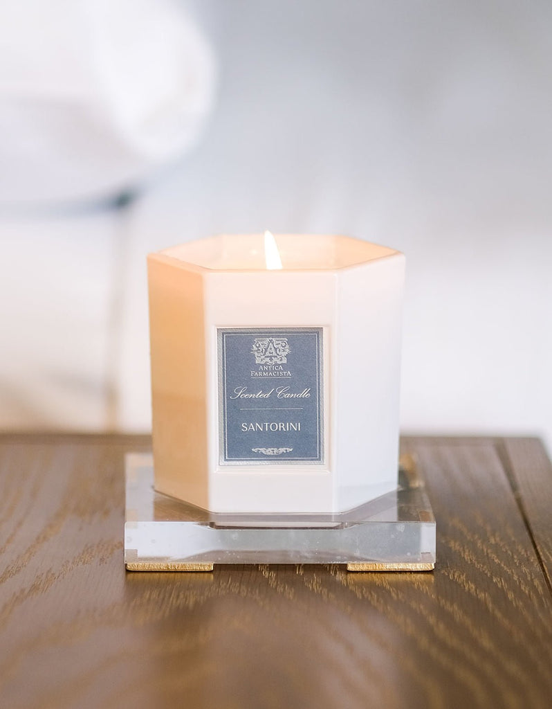 A lit, white octagonal 9oz candle labeled Santorini by Antica Farmacista sits on a clear acrylic tray from Antica Farmacista, placed on a wooden surface with a blurred background, emitting a warm glow.