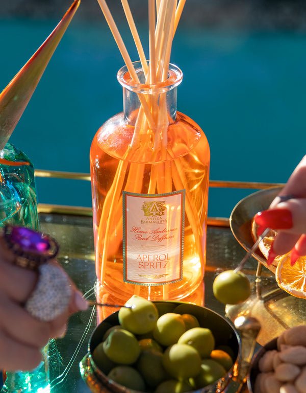 The Antica Farmacista - Aperol Spritz 250ml Reed Diffuser emits a citrus aroma. A hand with colorful rings reaches for green olives surrounded by snacks, against the backdrop of shimmering turquoise pool water.