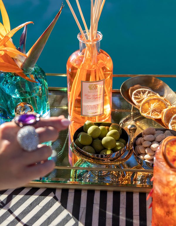 A sunny poolside scene features Antica Farmacistas Aperol Spritz 250ml Reed Diffuser beside a tray with olives, almonds, dried citrus slices, and vibrant flowers. A ring-adorned hand holds a cocktail above a tablecloth showcasing a black and white zigzag pattern that enhances the Citrus Spectrum theme.