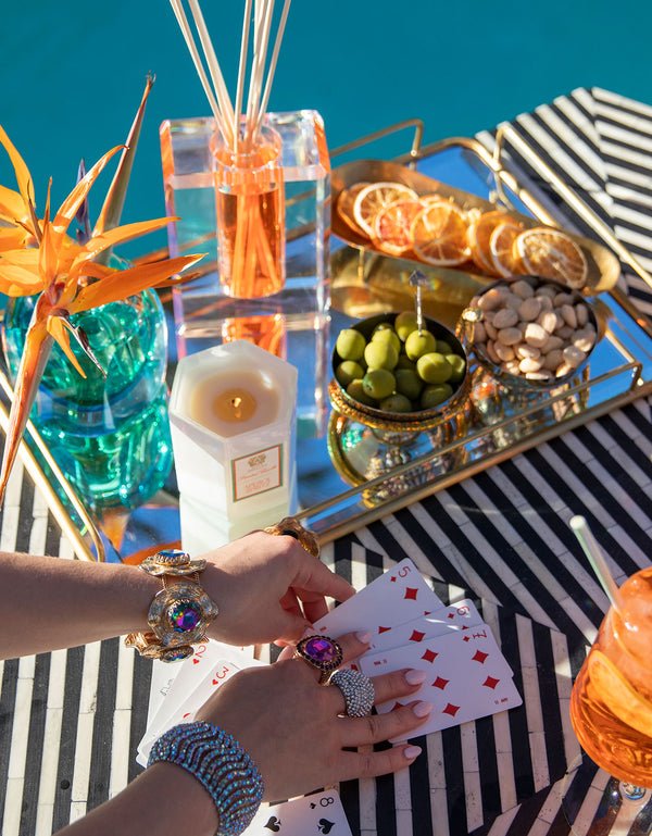 A colorful table by the poolside displays a hand holding playing cards. An Antica Farmacista - Aperol Spritz 9oz Candle flickers next to a bird of paradise flower vase, casting shadows on a tray of citrus slices, green olives, and nuts, creating a vibrant citrus spectrum.