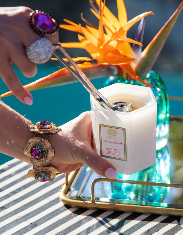 A hand with ornate rings and a bracelet lights an Antica Farmacista - Aperol Spritz 9oz Candle, made from a soy-paraffin wax blend, with a lighter and wick trimmer. The candle is on a gold tray near a blue vase holding bird of paradise flowers by the poolside.