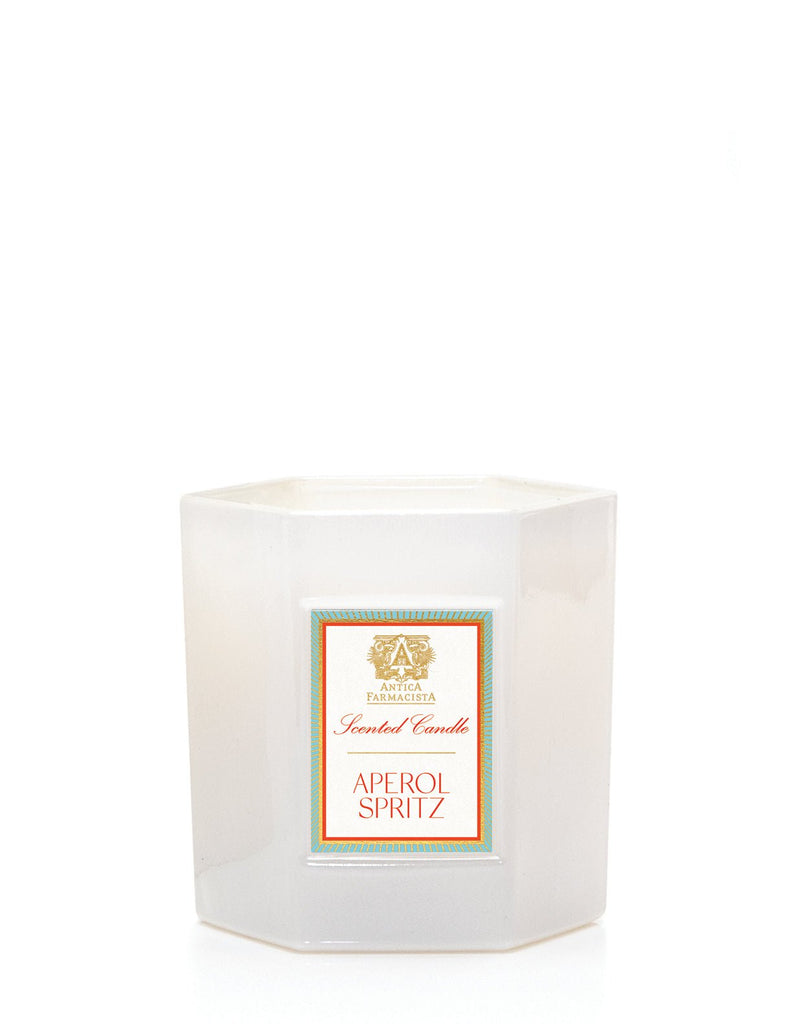 The Antica Farmacista Aperol Spritz 9oz Candle features a white hexagonal container with a label in gold and red text, framed by a turquoise border. Made from soy-paraffin wax, it promises to release the vibrant citrus aroma of an Aperol Spritz when lit.