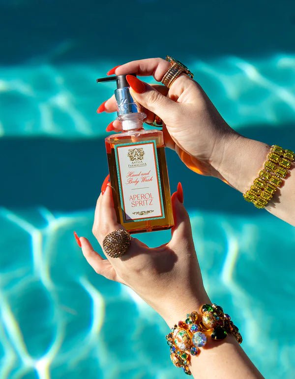 Hands adorned with colorful jewelry and red nails hold a bottle from the Antica Farmacista - Aperol Spritz Acrylic Bath & Body Gift Set against a sunlit pool, evoking a luxurious summer vibe. Infused with bitter orange essence, its perfect for any sunshine lover’s gift set.