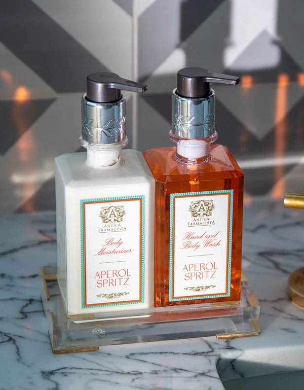 The Antica Farmacista - Aperol Spritz Acrylic Bath & Body Gift Set features a white body moisturizer and an orange hand and body wash with bitter orange essence, elegantly placed on a transparent tray on a marble surface under sunlit ambiance.