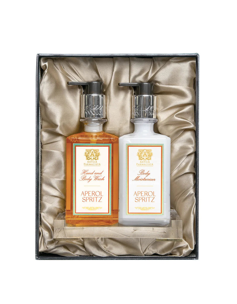 The Antica Farmacista - Aperol Spritz Acrylic Bath & Body Gift Set includes a scented body wash and moisturizer with Bitter Orange essence, both featuring pump dispensers. These elegantly rest on satin-like fabric inside the box.