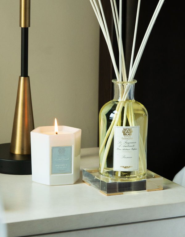 An Antica Farmacista Bergamot & Ocean Aira Candle in a white hexagonal holder is lit on a table beside a glass bottle of reed diffusers with oceanic notes, set on a clear square base. In the background, theres an elegant black and gold lamp.