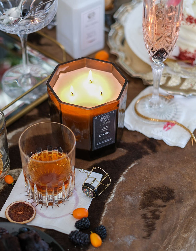 The Antica Farmacista Cask 3 Wick 25oz Candle emits a warm glow on a brown table, complemented by an amber drink with an orange slice, champagne flute, blackberries, small orange fruits, and a decorative napkin. The cozy and elegant ambiance is perfectly set.