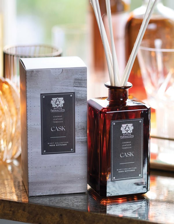 The Antica Farmacista - Cask 320ml Reed Diffuser from Antica Farmacista exudes an apothecary charm with its brown glass design and white reeds, elegantly set against a reflective surface and blurred glassware, complemented by its matching box.