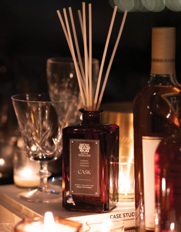 A dimly lit scene showcases Antica Farmacistas Cask 320ml Reed Diffuser with white sticks, exuding an apothecary vibe. Around it are empty wine glasses and a partially visible bottle, all enhanced by soft candlelight.