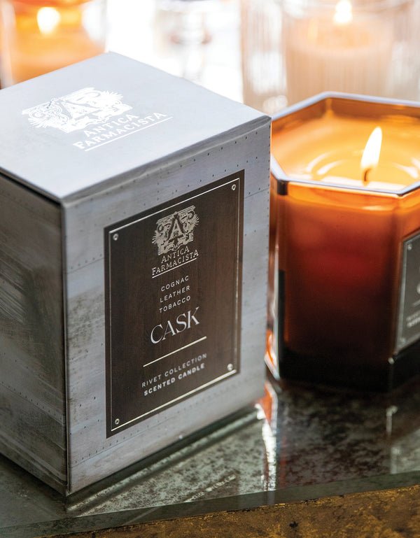 A close-up of the Cask 9oz Candle by Antica Farmacista in a brown jar beside its matching box, exuding rich notes of cognac, leather, and tobacco. The candles warm glow on a reflective surface fills the air with its captivating fragrance.