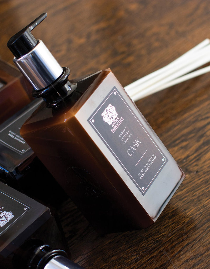 A brown bottle of Antica Farmacista - Cask Body Moisturizer 16oz with a pump dispenser lies on a wooden surface. The label features the Antica Farmacista logo and notes of rich cognac. Nearby, similar bottles hint at the soothing shea butter for a luxurious experience.