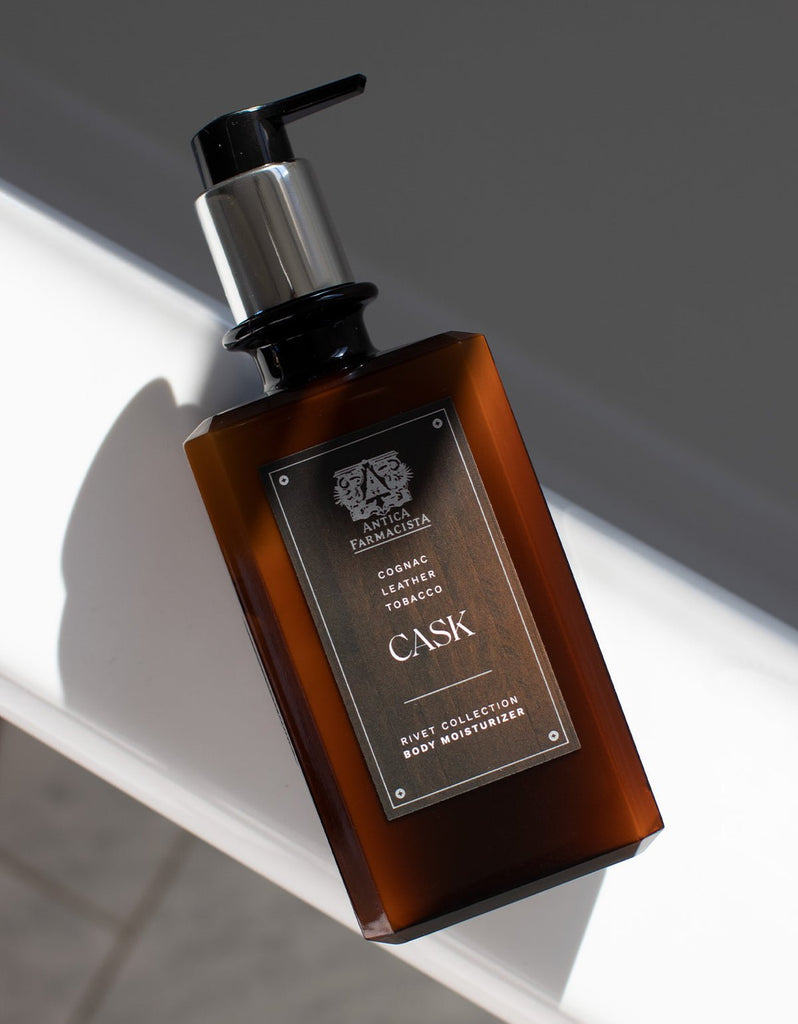 A dark amber bottle of Antica Farmacistas 16oz Cask Body Moisturizer, infused with Shea Butter, labeled Rich Cognac, Leather, Tobacco, features a black pump and metallic cap. It is elegantly placed on a white surface with shadows.