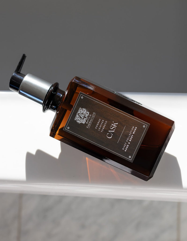 A rectangular amber bottle of Antica Farmacistas Cask Hand & Body Wash 16oz with a black pump sits on a white surface. The Antica Farmacista label features a dark design and cognac hints, casting shadows on the surface against a neutral gray background.