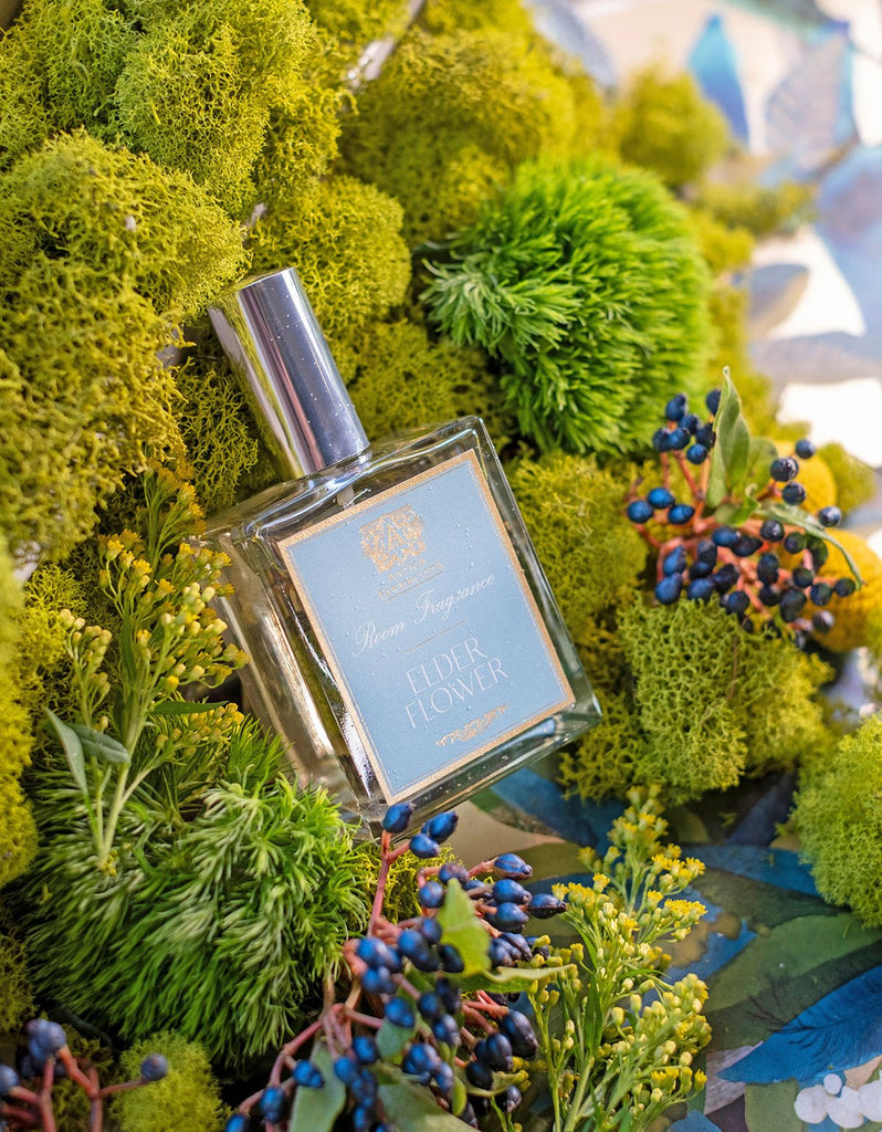 The Antica Farmacista - Elder Flower 100ml Room Spray rests amid vibrant moss and dark berries with yellow flowers, evoking botanical poetry against a blue and beige pattern backdrop, capturing nature’s serene elegance.