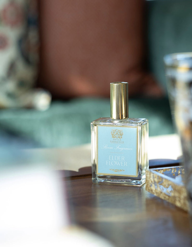 A square glass bottle of Antica Farmacistas Elder Flower 100ml Room Spray, topped with a golden cap, rests elegantly on a wooden surface. The softly blurred green and brown background suggests a chic interior that evokes botanical poetry.