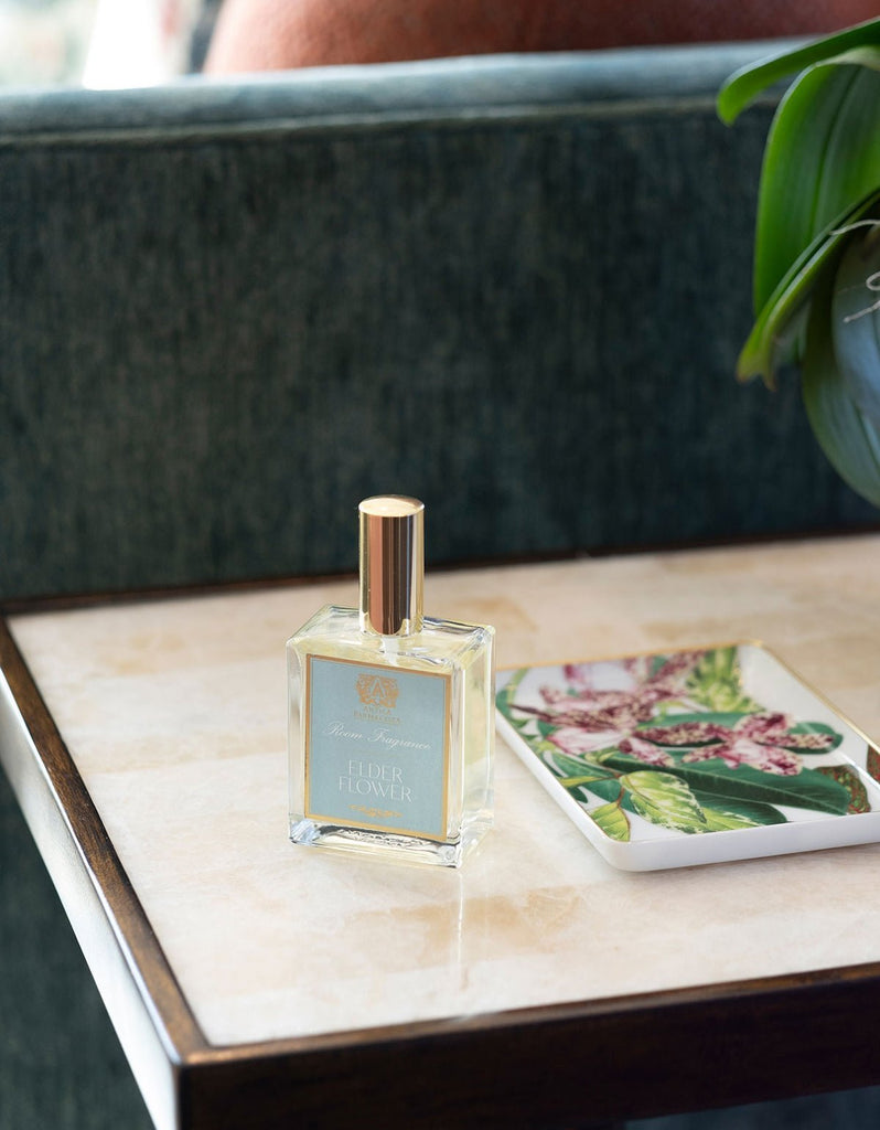 An Antica Farmacista Elder Flower 100ml Room Spray, capped in gold, rests on a marble table near a floral coaster. The setting exudes botanical poetry, complemented by the grace of a green velvet chair and the vibrant presence of greenery.