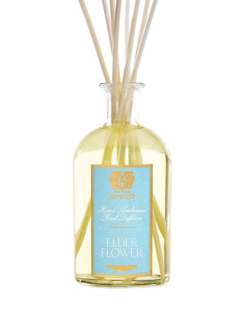 A 250ml Antica Farmacista glass bottle, labeled Elder Flower Reed Diffuser, contains a yellow liquid and several reed sticks. The blue label with gold and white text reads White Floral Home Ambiance, evoking a botanical fragrance.
.