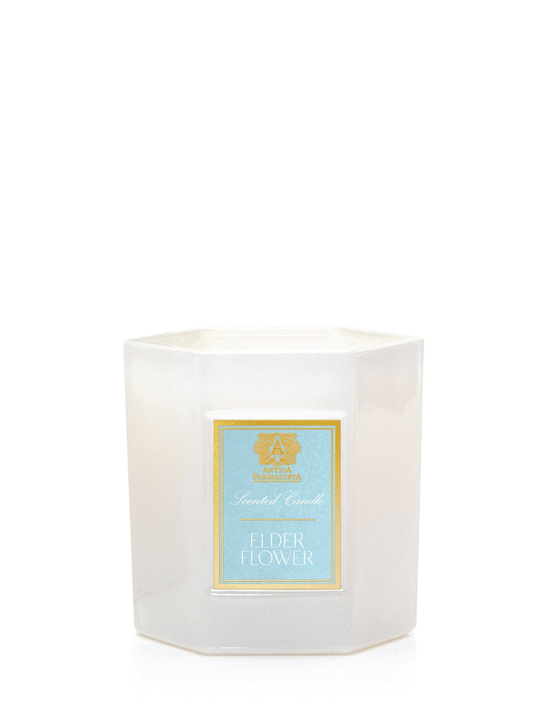 The Antica Farmacista Elder Flower candle features a white hexagonal jar with a light blue label reading Scented Candle. The bottom displays Elderflower amid gold decorations, enhancing its soy-paraffin wax blend with botanical elegance.