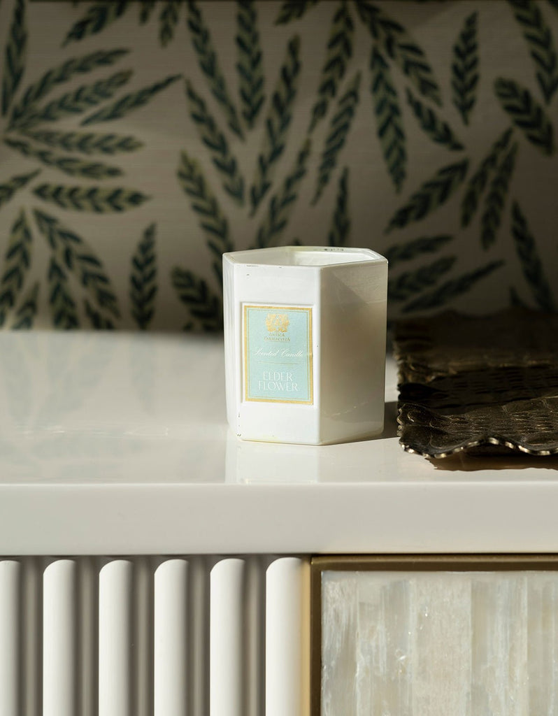 A white Antica Farmacista - Elder Flower 9oz Candle made from soy-paraffin wax rests on a cream shelf. The background showcases green leaf pattern wallpaper, evoking botanical poetry, with a woven brown mat partially visible on the right.