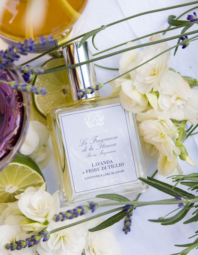 The Antica Farmacista - Lavender & Lime Blossom 100ml Room Spray bottle is surrounded by white flowers, lavender, lime blossoms, and a glass of purple liquid. A gold cap is partly visible at the top.