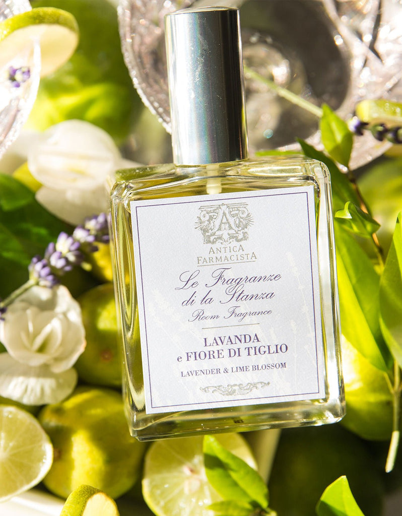 The Antica Farmacista Lavender & Lime Blossom 100ml Room Spray is elegantly displayed among fresh limes, green leaves, and white lime blossom, with sunlight enhancing the natural ambiance.