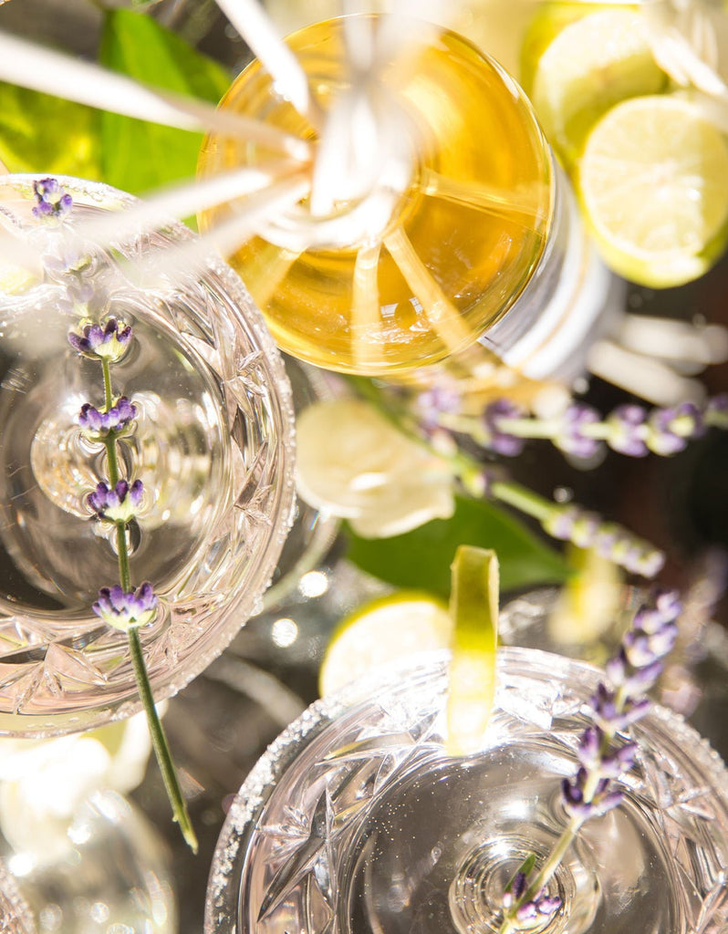 A close-up of a table featuring crystal glasses with yellow drinks garnished with lavender sprigs. Lemon slices, white flowers, and a touch of Antica Farmacistas Lavender & Lime Blossom reed diffuser mingle in the sunlight, casting highlights and shadows on the glass surfaces.