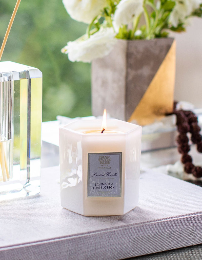 An Antica Farmacista - Lavender & Lime Blossom 9oz Candle in an octagonal white holder is placed on a gray cloth-covered book. A decorative vase with white flowers and a clear reed diffuser blur in the background.