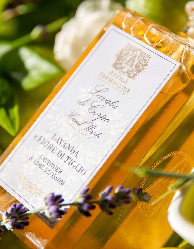 A close-up of Antica Farmacistas Lavender & Lime Blossom Hand & Body Wash, surrounded by lavender and greenery against a sunny backdrop. The amber liquid, crafted with natural ingredients, promises a rejuvenating cleanse.