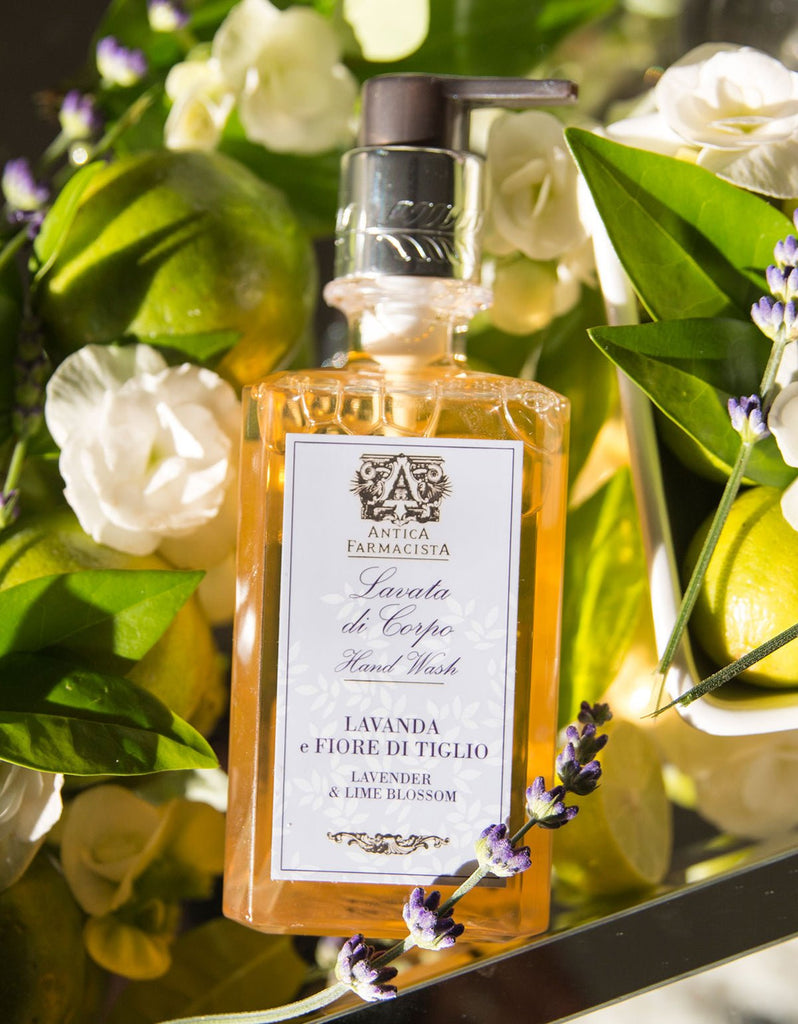 A bottle of Antica Farmacista - Lavender & Lime Blossom Hand & Body Wash 10oz is surrounded by white flowers, green leaves, and lavender in soft lighting that highlights the natural ingredients.