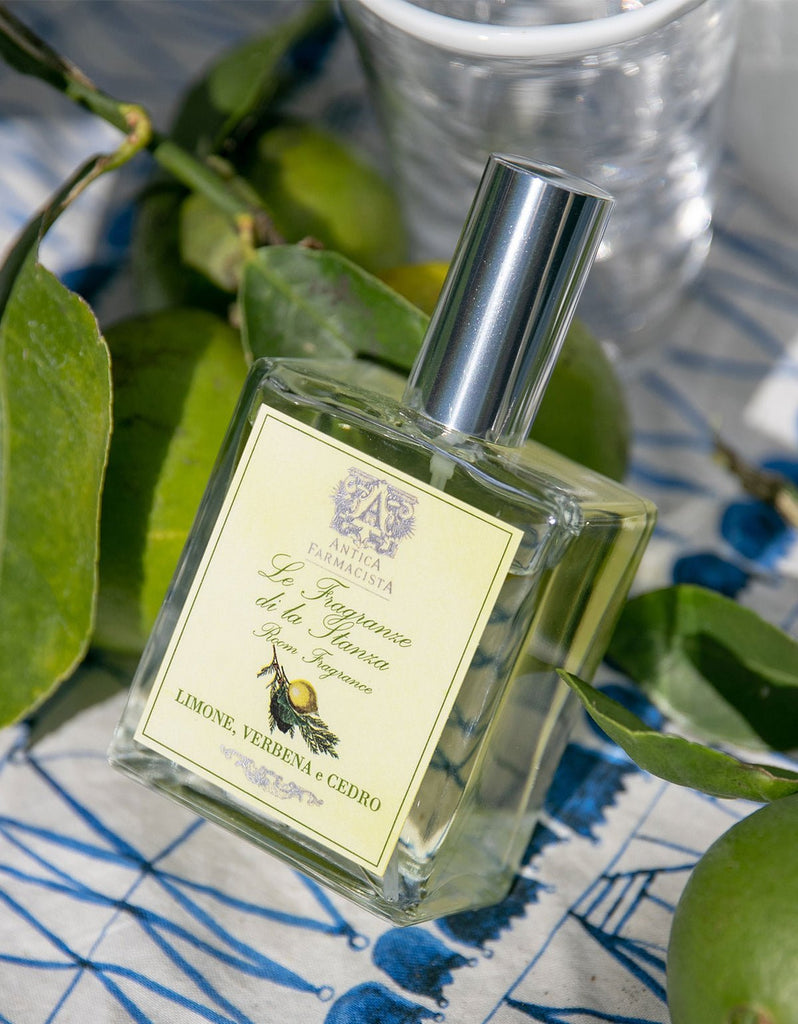 A bottle of Antica Farmacista - Lemon, Verbena & Cedar 100ml Room Spray rests on a patterned surface amid lush green leaves and lemons, exuding its fresh aroma. The vibrant setting enhances the refreshing scent.