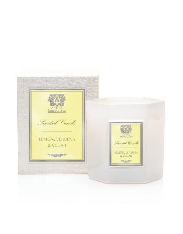 An Antica Farmacista - Lemon Verbena & Cedar Candle in a hexagonal glass holder is paired with its box, both showcasing the brands logo. The simple white background enhances the apothecary-style scene.